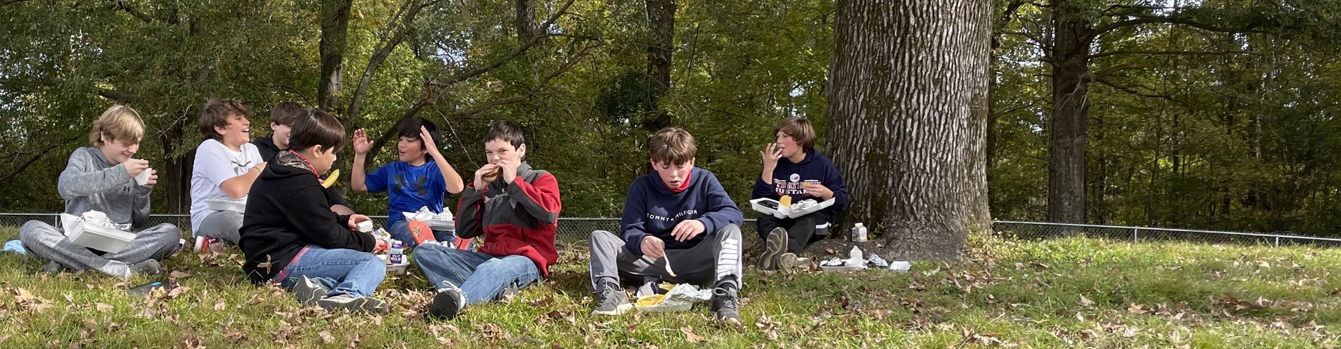 Boys eating lunch as a group on the grass under trees